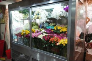 The image shows a commercial refrigerated floral display case filled with colorful fresh flowers including white, purple, yellow, and pink blooms arranged for sale in what appears to be a flower shop or market.