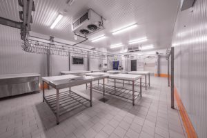 The image shows a sterile, white commercial kitchen or food processing facility with stainless steel work tables, overhead rail systems, and industrial refrigeration units mounted on the ceiling.