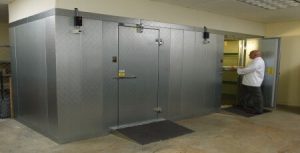 The image shows a large commercial walk-in freezer or cold storage unit with stainless steel doors and walls, with a person in white clothing working nearby in what appears to be a commercial kitchen or food service facility.