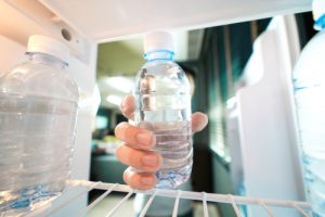 A person holds a clear plastic water bottle while standing in front of an open refrigerator.Retry