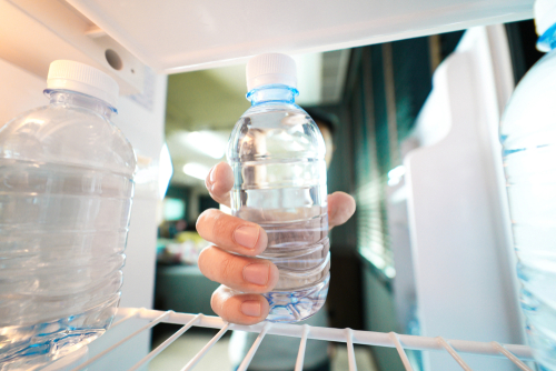 A person holds a clear plastic water bottle while standing in front of an open refrigerator.Retry